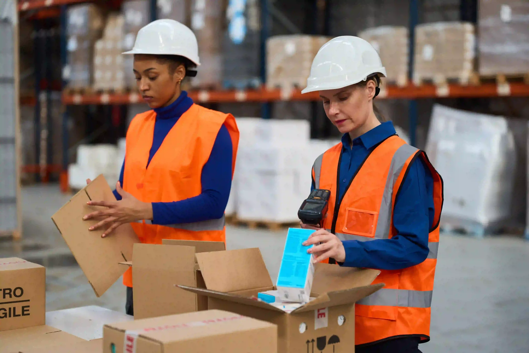Two female workers wearing safety vests and hard hats, packing and scanning products in a busy logistics warehouse